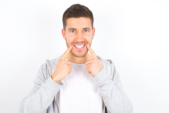 Strong healthy straight white teeth. Close up portrait of happy young caucasian man wearing casual clothes over white background  with beaming smile pointing on perfect clear white teeth.