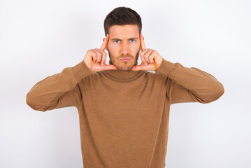 Serious concentrated young caucasian man wearing knitted turtleneck over white wall keeps fingers on temples, tries to ease tension, gather with thoughts and remember important information for exam