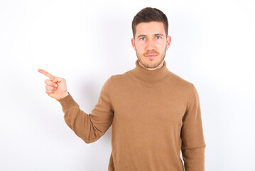 young caucasian man wearing knitted turtleneck over white background points to side on blank space demonstrates advertisement. People and promotion concept