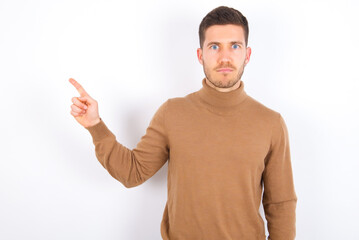 Serious young caucasian man wearing knitted turtleneck over white background smirks face points away on copy space shows something unpleasant. Look at this advertisement. Big price concept