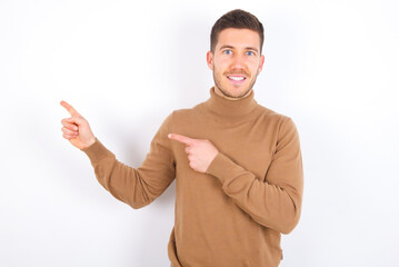 Optimistic young caucasian man wearing knitted turtleneck over white background points with both hands and  looking at empty space.