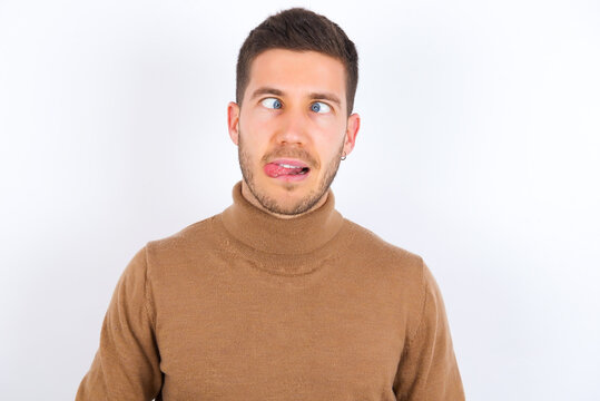 Funny young caucasian man wearing knitted turtleneck over white background makes grimace and crosses eyes plays fool has fun alone sticks out tongue.