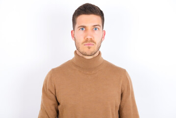 Displeased young caucasian man wearing knitted turtleneck over white background frowns face feels...