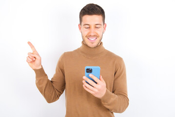 Smiling young caucasian man wearing knitted turtleneck over white background pointing finger at blank space holding phone in one hand