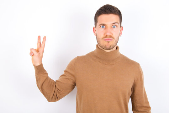 young caucasian man wearing knitted turtleneck over white background makes peace gesture keeps lips folded shows v sign. Body language concept