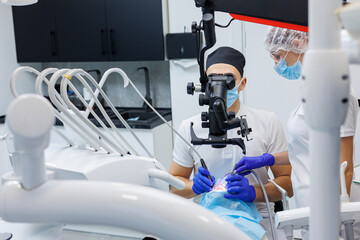 A successful dentist looks at the patient's teeth with a dental microscope and holds dental instruments near his mouth. The assistant helps the doctor. They wear white uniforms with masks and gloves.
