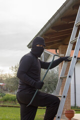 Thief dressed in black with crowbar and balaclava climbing a ladder while trying to sneak into a house