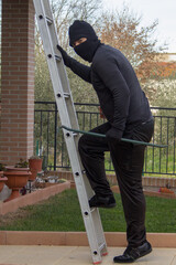 Thief dressed in black with crowbar and balaclava climbing a ladder while trying to sneak into a...