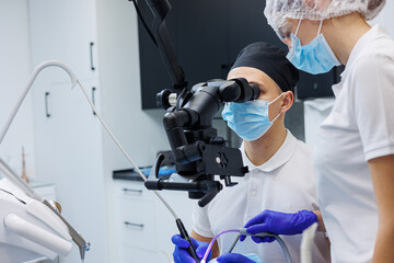 A successful dentist looks at the patient's teeth with a dental microscope and holds dental instruments near his mouth. The assistant helps the doctor. They wear white uniforms with masks and gloves.