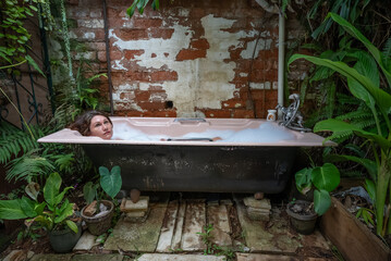 A young girl lying back in an outdoor bubble bath surrounded by lush vegetation. She is deep in...