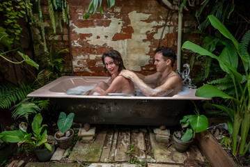 A couple enjoys a foam bath in the tropical outdoors. The young woman is glancing to the side and...