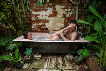 A young couple relaxes in an outdoor bubble bath among tropical plants. She playfully pushes her...