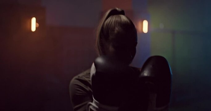 Portrait, a girl professionally practices punches in a dark hall of a fight club, boxing gloves on her hands. Silhouette, a girl boxer strikes in fighting gloves, a serious look of the face. Dark room