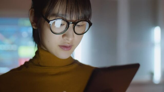 Arc shot of young Asian businesswoman in glasses using digital tablet while standing in dark office of investment company at night