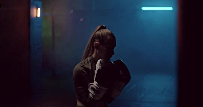 Silhouette, a woman boxer professionally makes strong punches on a punching bag in a dark hall of a fight club, preparing for a fight. Young female boxer practices punches in boxing gloves before a