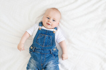 cute baby smiles lying on a white bed at home in a denim suit