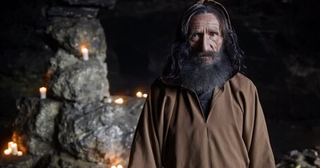 Portrait of a medieval bearded monk taking off the hood of his robe standing in a dark cave with candlelight