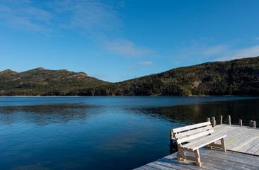 Obraz premium The view of two rolling hills covered in green trees from a wooden wharf with a bench on the deck of a wooden pier. The mountains are reflected in the still water. The sky is a vibrant blue with cloud