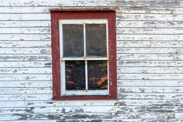 The exterior wall of a vintage white shed with a single hung closed window. The trim on the windows is a vibrant red. The white paint is peeling on the wood siding of the retro storage building. 