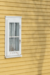An exterior yellow clapboard siding wooden wall with a single four pane closed window. There are circles on the bottom for ventilation. The shadow of a tall tree is projecting on the wall of the house
