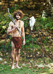Making a new shelter. A young caveman standing with wood in his hand and a partly-built hut behind him.