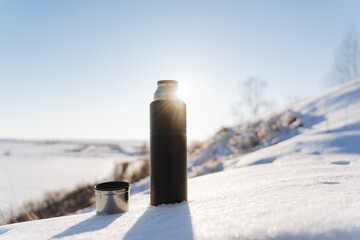Sunlight glints with rays against the blue sky. A thermos of hot tea stands in the snow. Winter landscape, hiking in the forest, camping equipment.