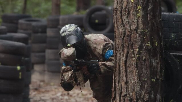 Play paintball game, players with guns. Painterly team in uniform and masks, extreme sport. Opposing teams of players shooting on paintball playing field outdoors. Man hides behind a tree trunk