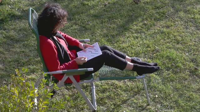 Brunette woman solving crossword puzzle in the garden. Woman dressed in a red shirt and black pants playing corssword. Home garden. Bright sunny day. Brain games. Memory training.
