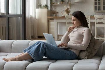 Obraz premium Pensive young woman sit on sofa staring at wireless computer screen feel concerned, looking thoughtful, working from home, search solution or ideas, choose goods online. Telecommute, challenge concept