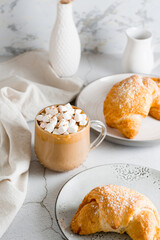 Cup of coffee with marshmallows and croissants on plates on the table. Homemade breakfast lifestyle. Vertical view