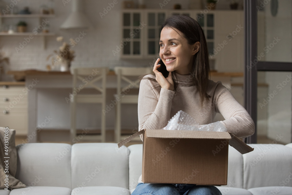 Wall mural smiling young woman sits on sofa at home with cardboard parcel, using smartphone talking to friend, 