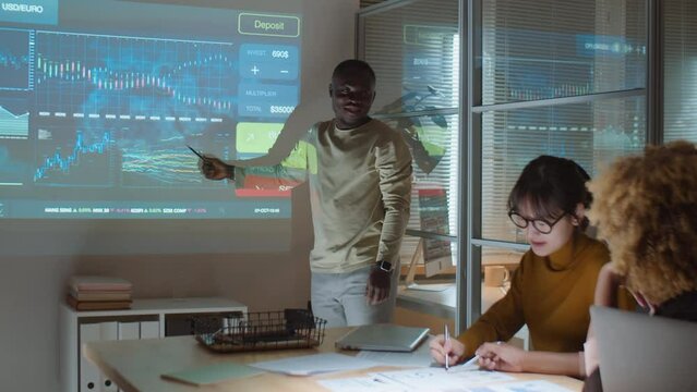 African American man showing stock market charts on projection screen and speaking with diverse female colleagues while working in team during night meeting in office of investment company