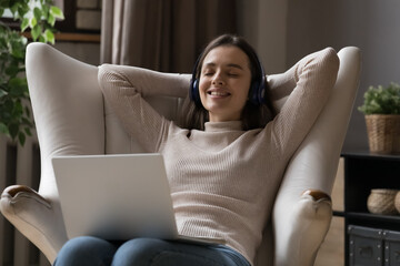 Serene woman put hands behind head leaned on armchair relax at home with laptop, enjoy track through wireless headphones spend carefree leisure use modern tech, digital streaming music service concept