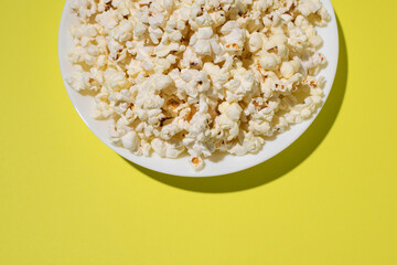 Popcorn on a yellow background. Delicious fresh homemade popcorn on a white plate. Close-up, selective focus. Top view. Cropped shot with hard shadows.
