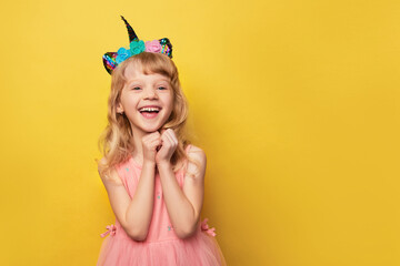Close-up portrait of a cute attractive cheerful girl. Cute smiling child wearing a festive unicorn headband. Blonde girl 5 years old in a pink dress on a bright yellow background with a place for text