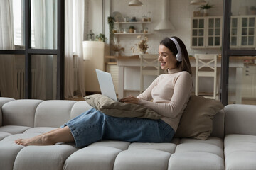 Smiling attractive young woman in headphones use laptop seated on cozy sofa in living room. Makes...