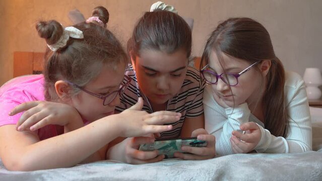 Three little female friends were lying on the bed ,talking and laughing.They use a smart phone to play games.	
