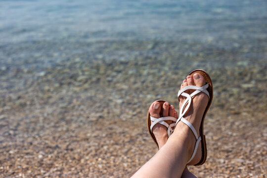 Women's legs in white sandals on the background of turquoise water on a pebble beach