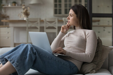 Thoughtful woman sit on couch with laptop staring away, looks pensive, consider online purchase,...