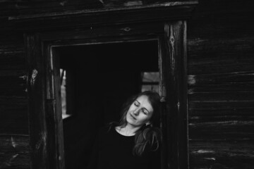 Fototapeta na wymiar beautiful Ukrainian girl in black clothes near the old wooden house. The war in Ukraine. Portrait of a woman on a dark wooden background. Old abandoned wooden house. Old wooden window frame