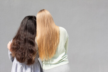 two girls with different color of hair are standing and hugging each other, grey background wall, rear view
