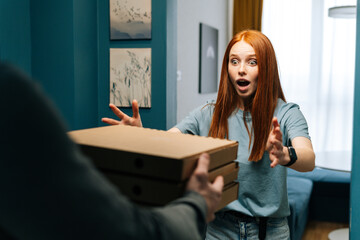 Surprised young woman receiving paper boxes with hot pizza from unrecognizable courier male on...