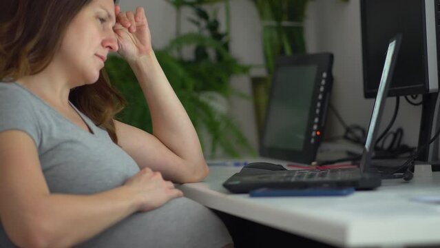 A pregnant woman is working on a laptop. Tired girl, increased work fatigue. Businesswoman typing on a laptop while sitting at her workplace in the office. Work during pregnancy from home.