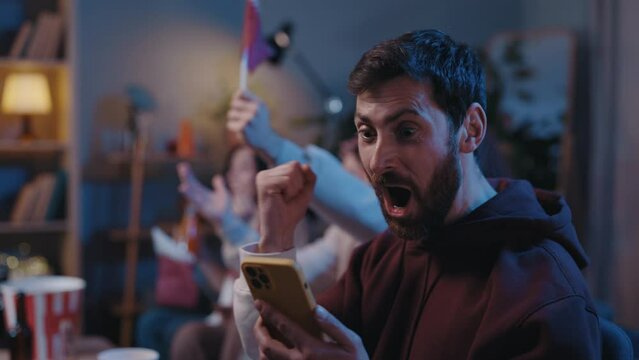 Concentrated bearded man looking attentively at his device screen and rejoicing while looking at results of sports betting during football game. Male fans cheering at home concept.