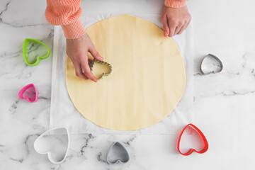 Hands of caucasian teenage girl cut dough with metal heart cutter on white parchment.
