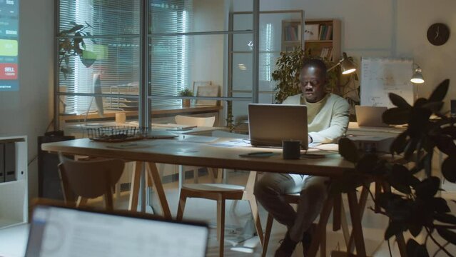 Wide shot of African American businessman working on laptop at desk in dark office during night