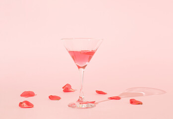 Cocktail glass and red flower petals with shadow on pink background. MInimal spring or season party concept.