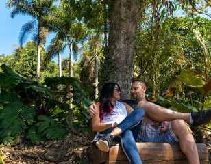 Naklejka na ściany i meble Casal sentando em banco de madeira ornamental, em visita ao museu a céu aberto em Minas Gerais, meio a muita vegetação, grandes árvores e lindos coqueiros.
