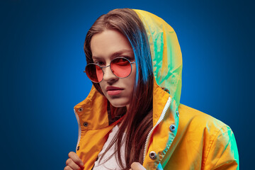 Portrait of fashion young girl in yellow raincoat and round sunglasses in neon light on blue...