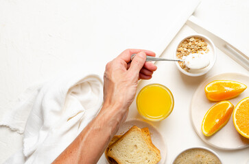 A cup of coffee, orange juice, some toasts, pieces of orange, and a hand taking a spoonful of plain yoghurt with granola in a white tray with a white napkin, on a white background. 